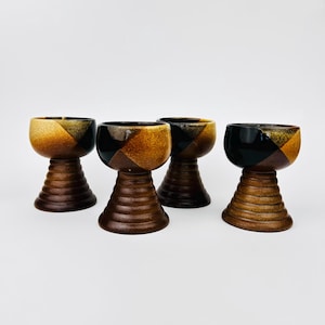 Puede incluir: Juego de cuatro vasos para beber de cerámica con un patrón geométrico en tonos marrones, negros y amarillos. Los vasos tienen una copa acampanada y un tallo corto y cónico.