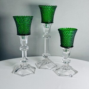 K&ouml;nnte beinhalten: Ein Set aus drei Kerzenhaltern mit klaren, facettierten Glassch&auml;ften und sechseckigen F&uuml;&szlig;en. Jeder Halter ist mit einem leuchtend smaragdgr&uuml;nen Glasbecher versehen, der eine texturierte Noppenstruktur auf der gesamten Au&szlig;enfl&auml;che aufweist.
