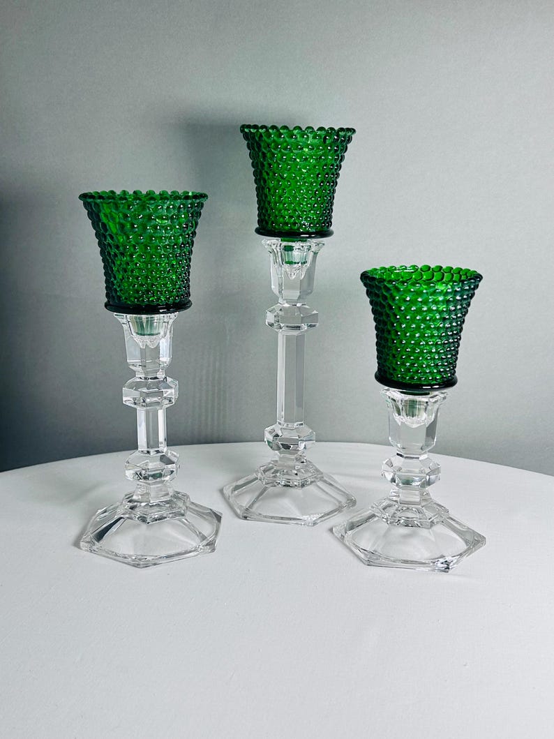 K&ouml;nnte beinhalten: Ein Set aus drei Kerzenhaltern mit klaren, facettierten Glassch&auml;ften und sechseckigen F&uuml;&szlig;en. Jeder Halter ist mit einem leuchtend smaragdgr&uuml;nen Glasbecher versehen, der eine texturierte Noppenstruktur auf der gesamten Au&szlig;enfl&auml;che aufweist.