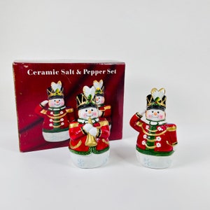 May include: Ceramic salt and pepper shaker set featuring three snowman figurines dressed as toy soldiers. The snowmen are wearing red and green uniforms with gold accents. The box reads "Ceramic Salt & Pepper Set".