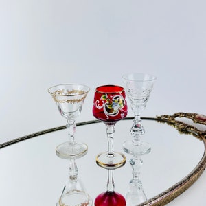 May include: Three vintage glass shot glasses on a mirrored tray. One glass is clear with a cut design, one is red with gold and white floral designs, and one is clear with a gold rim.