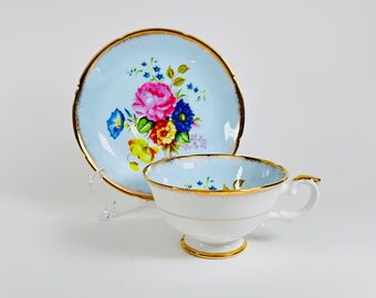 Può includere: Un delicato set da tè composto da tazza e piattino con un motivo floreale. Il piattino è azzurro con un bordo dorato e presenta un bouquet di fiori rosa, gialli, blu e arancioni. La tazza da tè è bianca con un bordo dorato e un motivo floreale abbinato.