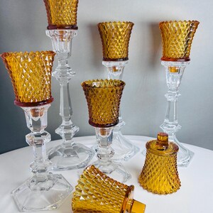 K&ouml;nnte beinhalten: Eine Sammlung von Kerzenhaltern aus klarem Glas mit bernsteinfarbenen Glaseins&auml;tzen im Rautenmuster. Das Set besteht aus mehreren hohen, facettierten St&auml;ndern aus klarem Glas und mehreren abnehmbaren bernsteinfarbenen Glaseins&auml;tzen mit Textur.