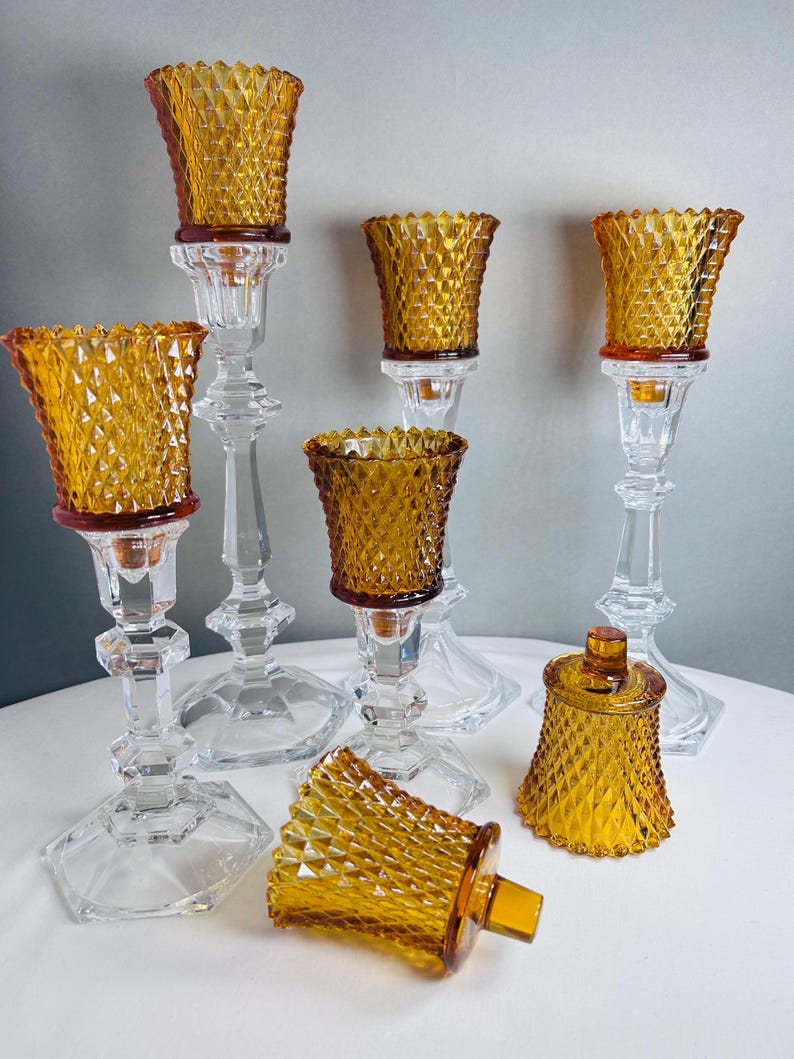 K&ouml;nnte beinhalten: Eine Sammlung von Kerzenhaltern aus klarem Glas mit bernsteinfarbenen Glaseins&auml;tzen im Rautenmuster. Das Set besteht aus mehreren hohen, facettierten St&auml;ndern aus klarem Glas und mehreren abnehmbaren bernsteinfarbenen Glaseins&auml;tzen mit Textur.