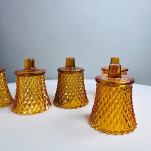 K&ouml;nnte beinhalten: Ein Set aus f&uuml;nf bernsteinfarbenen Votivkerzenhaltern aus Glas, die jeweils eine ausgepr&auml;gte Rautenstruktur auf der Au&szlig;enseite und einen kleinen, zylindrischen Knauf auf dem Deckel aufweisen und in einer Gruppe angeordnet sind.