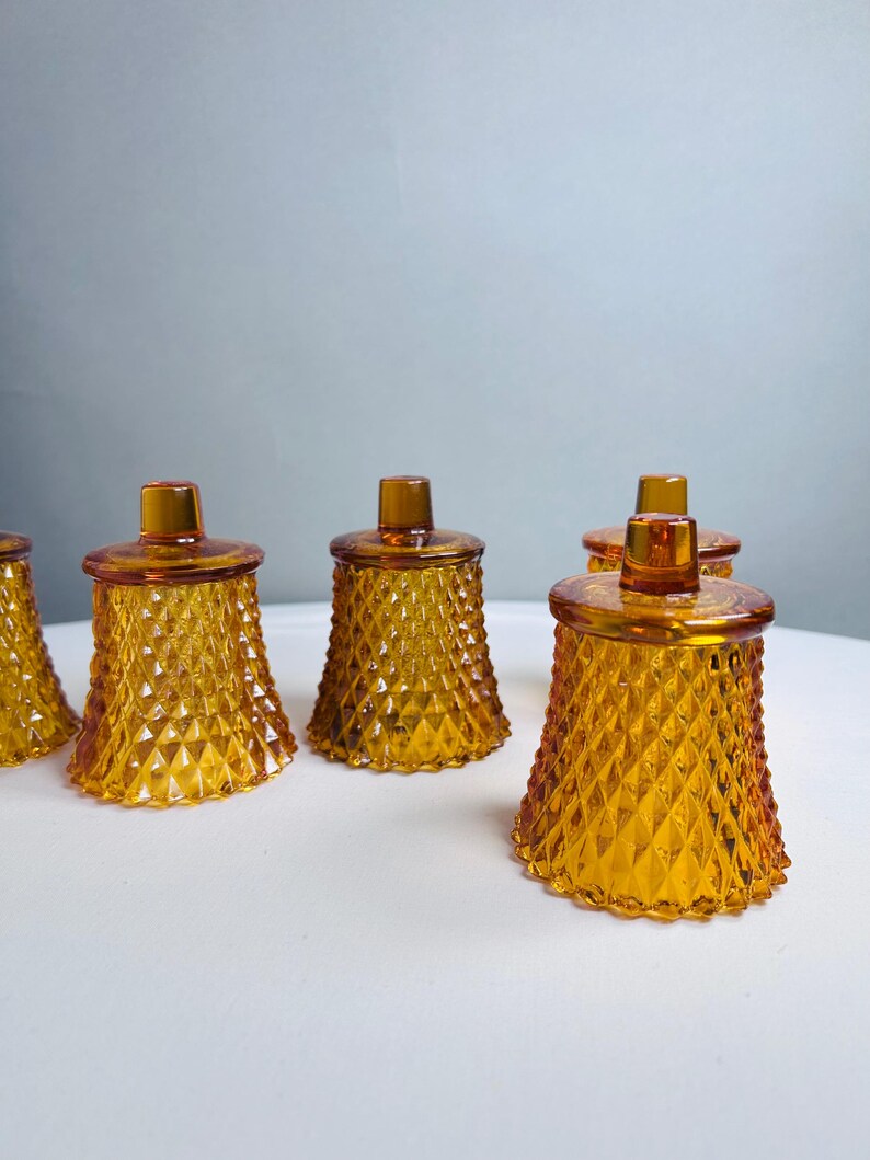 K&ouml;nnte beinhalten: Ein Set aus f&uuml;nf bernsteinfarbenen Votivkerzenhaltern aus Glas, die jeweils eine ausgepr&auml;gte Rautenstruktur auf der Au&szlig;enseite und einen kleinen, zylindrischen Knauf auf dem Deckel aufweisen und in einer Gruppe angeordnet sind.