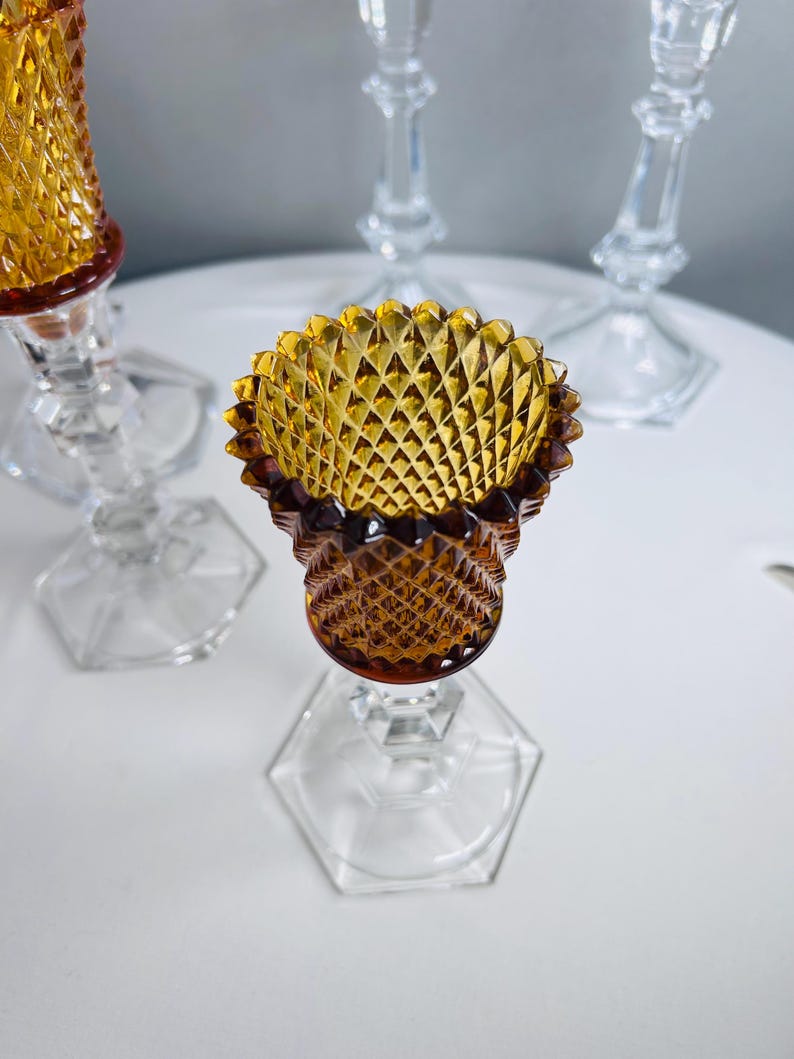 K&ouml;nnte beinhalten: Ein Kerzenhalter aus Glas mit einem bernsteinfarbenen, rautenf&ouml;rmig gemusterten Oberteil mit gewelltem Rand, das auf einem klaren, sechseckigen Glasfu&szlig; ruht. Das Design umfasst einen facettierten Stiel, der die bernsteinfarbene Schale verbindet.