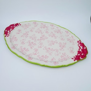 Puede incluir: Bandeja ovalada blanca con un patrón floral rosa y un borde festoneado verde. La bandeja tiene asas rosas con un diseño decorativo.