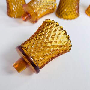 K&ouml;nnte beinhalten: Eine Sammlung von bernsteinfarbenen Votivkerzenhaltern aus Glas mit einem erhabenen Rautenmuster auf der Au&szlig;enfl&auml;che. Jeder Halter hat einen ausgestellten oberen Rand und einen runden Boden mit einem kleinen zentralen Vorsprung.