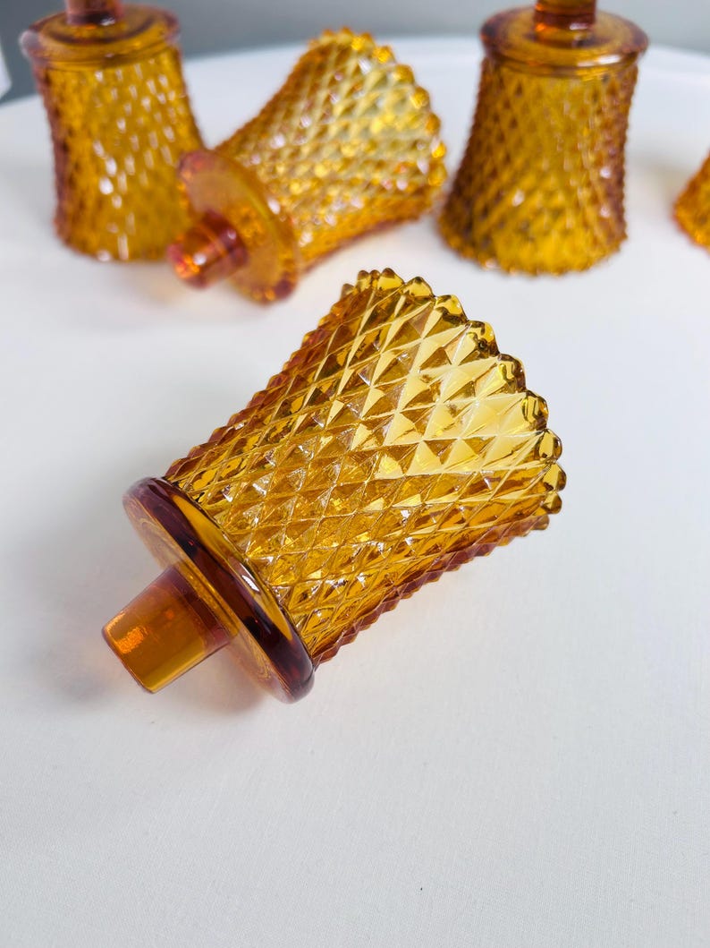 K&ouml;nnte beinhalten: Eine Sammlung von bernsteinfarbenen Votivkerzenhaltern aus Glas mit einem erhabenen Rautenmuster auf der Au&szlig;enfl&auml;che. Jeder Halter hat einen ausgestellten oberen Rand und einen runden Boden mit einem kleinen zentralen Vorsprung.