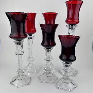 May include: Six glass candle holders with clear, faceted bases and stems. The tops are a deep red colour with a ruffled edge. The candle holders are arranged on a white surface, creating a striking display.