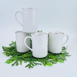 May include: Four white ceramic mugs with snowflake designs and gold rims are arranged on a bed of green foliage. The mugs have a classic handle design and a textured snowflake pattern, perfect for holiday beverages.