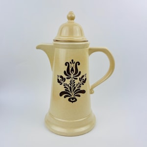 May include: A beige ceramic coffee pot with a brown floral design. The pot has a lid and a handle.