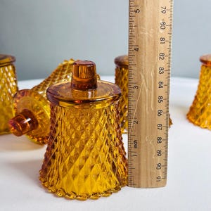 K&ouml;nnte beinhalten: Ein Set aus bernsteinfarbenen Glaskerzenhaltern mit Rautenmuster. Jeder Halter hat einen kleinen Knopf an der Oberseite und einen gewellten Fu&szlig;. Ein Holzlineal steht vertikal daneben und zeigt eine H&ouml;he von etwa 8,9 cm an.
