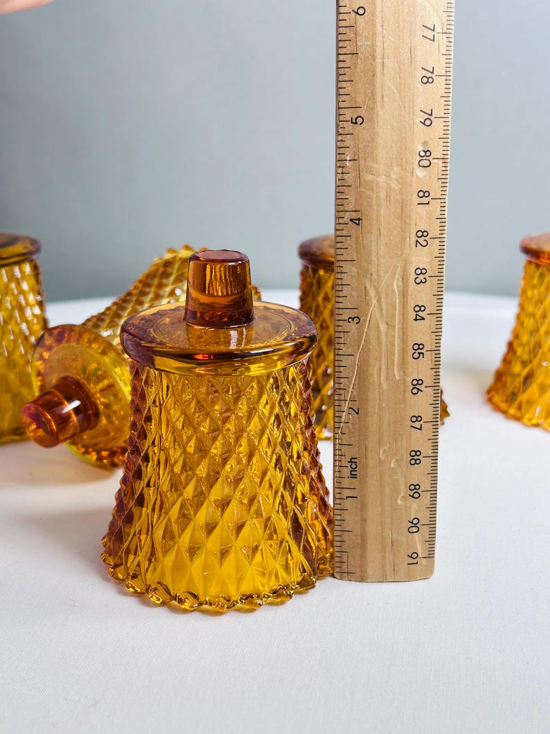K&ouml;nnte beinhalten: Ein Set aus bernsteinfarbenen Glaskerzenhaltern mit Rautenmuster. Jeder Halter hat einen kleinen Knopf an der Oberseite und einen gewellten Fu&szlig;. Ein Holzlineal steht vertikal daneben und zeigt eine H&ouml;he von etwa 8,9 cm an.