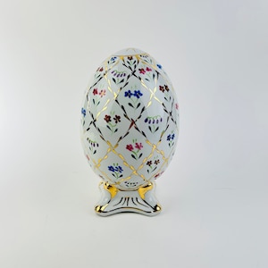 Könnte beinhalten: Ein weißes Porzellan-Ei mit goldenem und floralem Design. Das Ei ist mit einem Rautenmuster und kleinen Blumen in Rot-, Blau- und Lilatönen verziert. Das Ei steht auf einem goldenen Ständer mit drei Füßen.