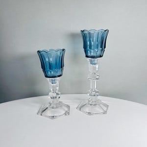 K&ouml;nnte beinhalten: Zwei Kerzenhalter aus Glas in unterschiedlichen H&ouml;hen, jeweils mit einer tiefblauen, gewellten Schale auf einem klaren, facettierten, sechseckigen Fu&szlig; und Stiel. Die Stiele haben ein mehrstufiges, geometrisches Design.