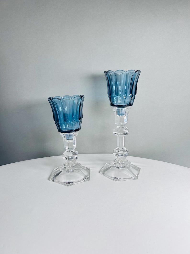 K&ouml;nnte beinhalten: Zwei Kerzenhalter aus Glas in unterschiedlichen H&ouml;hen, jeweils mit einer tiefblauen, gewellten Schale auf einem klaren, facettierten, sechseckigen Fu&szlig; und Stiel. Die Stiele haben ein mehrstufiges, geometrisches Design.