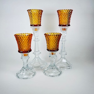 K&ouml;nnte beinhalten: Ein Set aus vier Kerzenhaltern aus Glas mit bernsteinfarbenen, rautenf&ouml;rmig gemusterten Kelchen, die auf klaren, facettierten Stielen und runden F&uuml;&szlig;en stehen. Die zwei h&ouml;heren Halter stehen hinter den zwei k&uuml;rzeren Kerzenhaltern.
