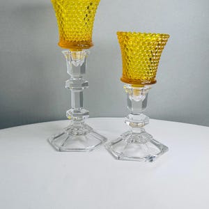 K&ouml;nnte beinhalten: Zwei Kerzenhalter mit klaren, facettierten Glasstielen, sechseckigen F&uuml;&szlig;en und abnehmbaren, leuchtend gelben Hobnail-Glaseins&auml;tzen. Die Glaseins&auml;tze weisen eine texturierte Oberfl&auml;che mit erhabenen Punkten auf, die einen Kontrast zum glatten, geometrischen Design der transparenten Glasst&auml;nder bildet.