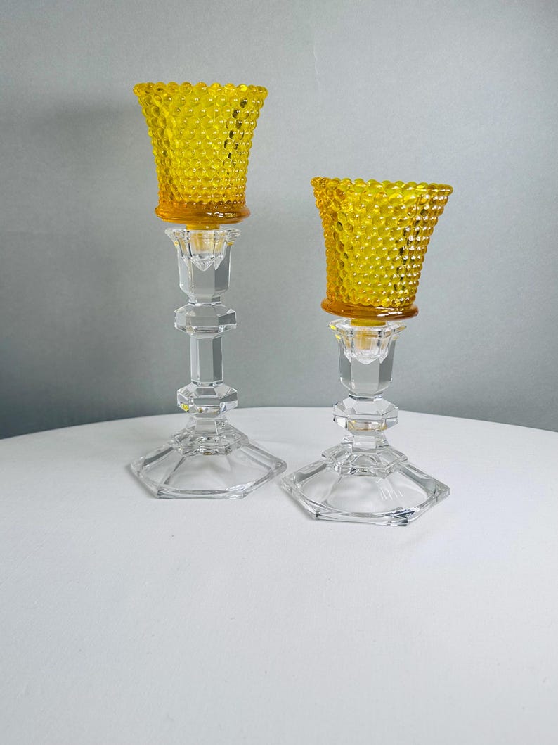 K&ouml;nnte beinhalten: Zwei Kerzenhalter mit klaren, facettierten Glasstielen, sechseckigen F&uuml;&szlig;en und abnehmbaren, leuchtend gelben Hobnail-Glaseins&auml;tzen. Die Glaseins&auml;tze weisen eine texturierte Oberfl&auml;che mit erhabenen Punkten auf, die einen Kontrast zum glatten, geometrischen Design der transparenten Glasst&auml;nder bildet.