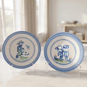 May include: Two round ceramic plates featuring hand-painted blue and green illustrations. The left plate depicts a figure with a hat and a small animal, while the right plate shows a stylized sheep. Both plates have blue circular rim accents.