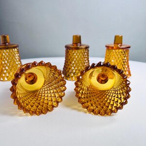 K&ouml;nnte beinhalten: Ein Set aus f&uuml;nf bernsteinfarbenen Votivkerzenhaltern aus Glas mit einem strukturierten Rautenmuster auf der Au&szlig;enseite. Das Glas ist durchscheinend und jeder Halter hat einen gewellten Rand sowie einen kleinen zylindrischen Vorsprung am Boden.