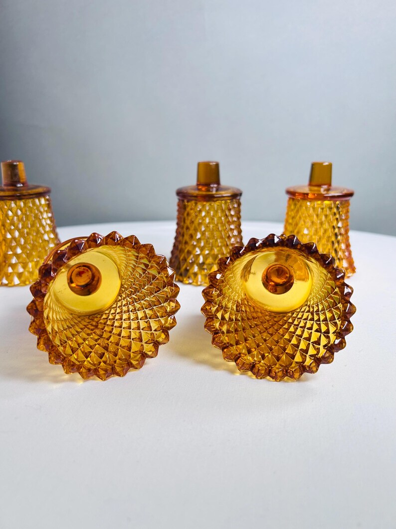 K&ouml;nnte beinhalten: Ein Set aus f&uuml;nf bernsteinfarbenen Votivkerzenhaltern aus Glas mit einem strukturierten Rautenmuster auf der Au&szlig;enseite. Das Glas ist durchscheinend und jeder Halter hat einen gewellten Rand sowie einen kleinen zylindrischen Vorsprung am Boden.