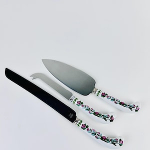 May include: A trio of serving implements: a cake server, a cheese knife, and a cake knife. The handles are white with a floral pattern in pink, green, and purple. The blades are silver and black.