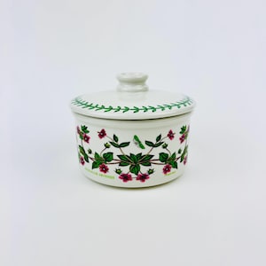 Puede incluir: Tarro de cerámica blanco con un diseño floral verde y rosa. El tarro tiene una tapa y el texto "Galium Aparine" está impreso en la parte inferior.