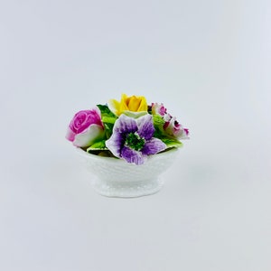 Puede incluir: Un arreglo floral de porcelana blanca con flores rosas, amarillas y moradas en una cesta blanca.