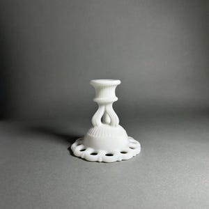 以下が含まれることがあります： 白のヴィンテージスタイルのキャンドルホルダー。ベースは丸く、スカラップエッジと装飾的な切り欠きがあります。ステムは砂時計型で、詳細な上部が付いています。キャンドルホルダーは不透明な白いガラス製です。