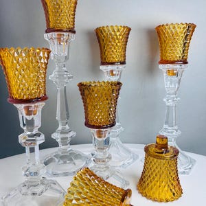 K&ouml;nnte beinhalten: Eine Sammlung von bernsteinfarbenen Glaskerzenhaltern mit Rautenmuster. Das Set umfasst mehrere hohe, klare Glasst&auml;nder mit bernsteinfarbenen Eins&auml;tzen sowie zwei kleinere bernsteinfarbene Glasgef&auml;&szlig;e, eines mit Deckel und eines auf der Seite liegend.