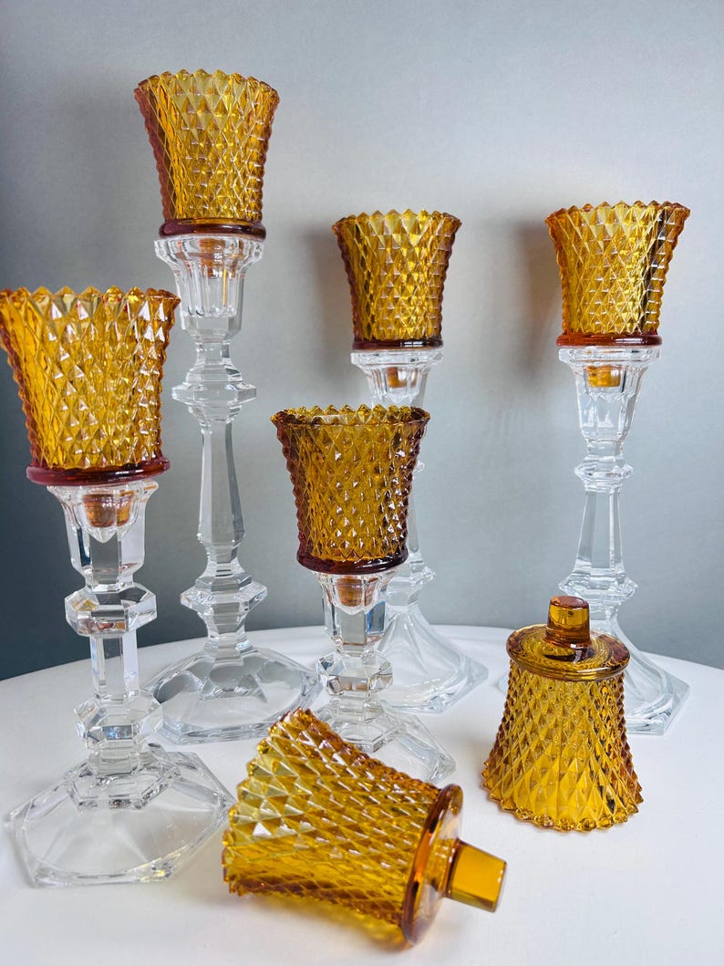 K&ouml;nnte beinhalten: Eine Sammlung von bernsteinfarbenen Glaskerzenhaltern mit Rautenmuster. Das Set umfasst mehrere hohe, klare Glasst&auml;nder mit bernsteinfarbenen Eins&auml;tzen sowie zwei kleinere bernsteinfarbene Glasgef&auml;&szlig;e, eines mit Deckel und eines auf der Seite liegend.