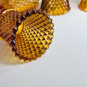 K&ouml;nnte beinhalten: Eine Sammlung von bernsteinfarbenen Glaskerzenhaltern mit einem ausgepr&auml;gten Rautenmuster auf der Au&szlig;enfl&auml;che. Jedes St&uuml;ck hat einen ausgestellten, gewellten Rand und eine durchscheinende Oberfl&auml;che, die das Licht einf&auml;ngt und die geometrische Textur hervorhebt.