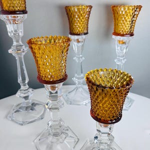 K&ouml;nnte beinhalten: Ein Set aus f&uuml;nf Kerzenhaltern mit klaren, facettierten Glassch&auml;ften auf sechseckigen F&uuml;&szlig;en und bernsteinfarbenen Glaskelchen mit Rautenmuster. Das Set umfasst verschiedene H&ouml;hen, jeweils mit einem strukturierten Votivglaseinsatz.