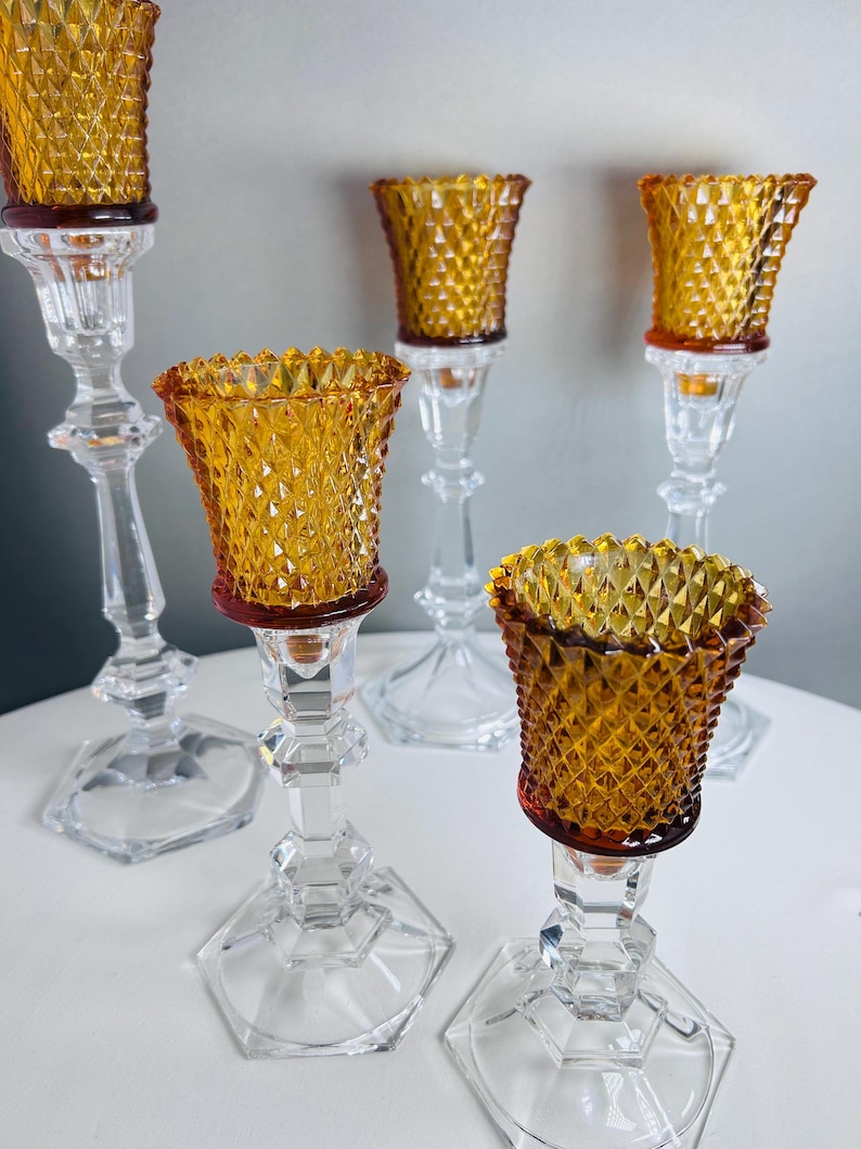K&ouml;nnte beinhalten: Ein Set aus f&uuml;nf Kerzenhaltern mit klaren, facettierten Glassch&auml;ften auf sechseckigen F&uuml;&szlig;en und bernsteinfarbenen Glaskelchen mit Rautenmuster. Das Set umfasst verschiedene H&ouml;hen, jeweils mit einem strukturierten Votivglaseinsatz.
