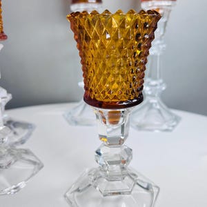K&ouml;nnte beinhalten: Ein Kerzenhalter mit einem bernsteinfarbenen Glasaufsatz, der ein Rautenmuster und einen gewellten Rand aufweist. Der Fu&szlig; besteht aus klarem, facettiertem Glas mit sechseckiger Basis und tr&auml;gt den bernsteinfarbenen Kelch &uuml;ber einen Stiel.