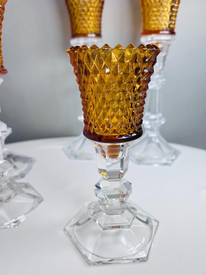 K&ouml;nnte beinhalten: Ein Kerzenhalter mit einem bernsteinfarbenen Glasaufsatz, der ein Rautenmuster und einen gewellten Rand aufweist. Der Fu&szlig; besteht aus klarem, facettiertem Glas mit sechseckiger Basis und tr&auml;gt den bernsteinfarbenen Kelch &uuml;ber einen Stiel.