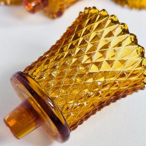 K&ouml;nnte beinhalten: Ein Votivkerzenhalter aus durchscheinendem bernsteinfarbenem Glas mit einem erhabenen Rautenmuster auf der Au&szlig;enfl&auml;che. Das Gef&auml;&szlig; hat einen breiten, gewellten Rand und einen runden Boden mit einem mittig hervorstehenden zylindrischen Glaszapfen.