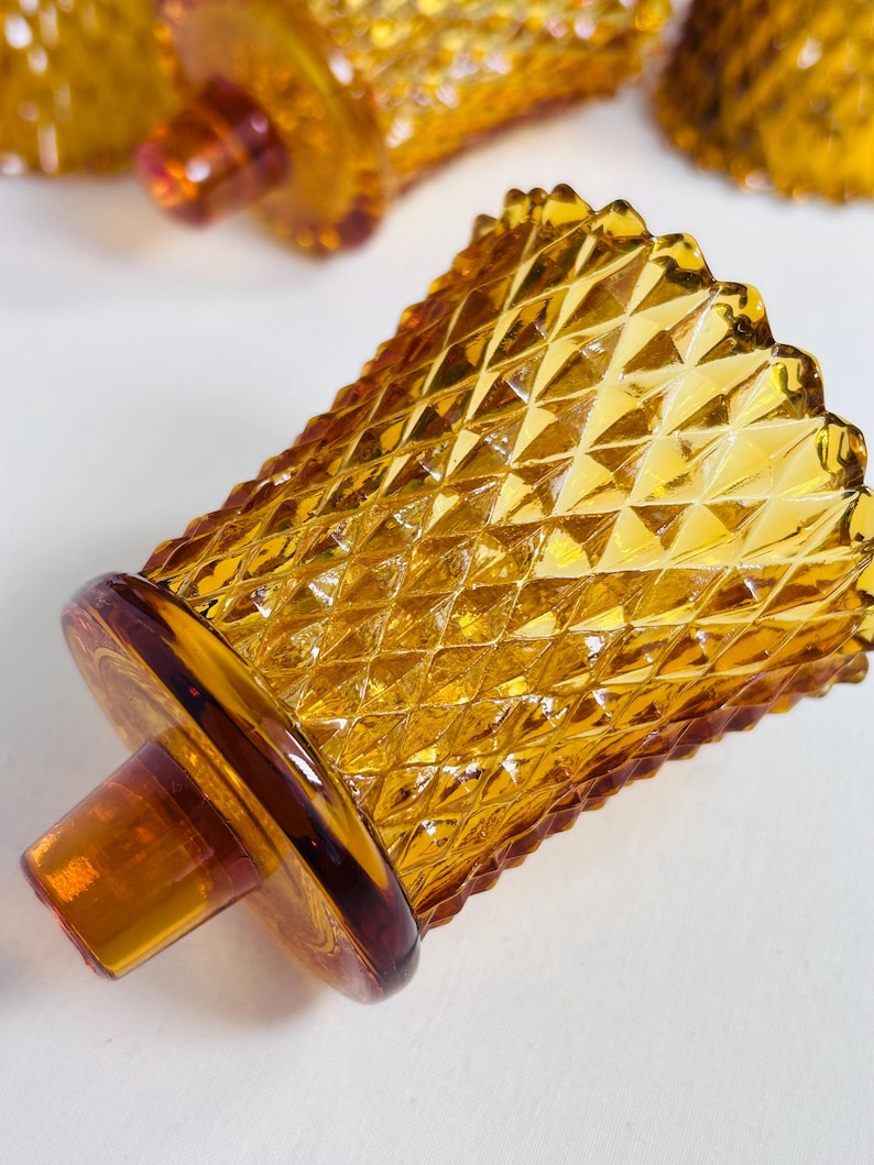 K&ouml;nnte beinhalten: Ein Votivkerzenhalter aus durchscheinendem bernsteinfarbenem Glas mit einem erhabenen Rautenmuster auf der Au&szlig;enfl&auml;che. Das Gef&auml;&szlig; hat einen breiten, gewellten Rand und einen runden Boden mit einem mittig hervorstehenden zylindrischen Glaszapfen.