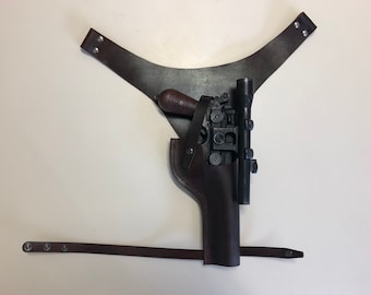 May include: A brown leather holster with a black gun and a scope attached. The holster has a strap that goes over the shoulder.