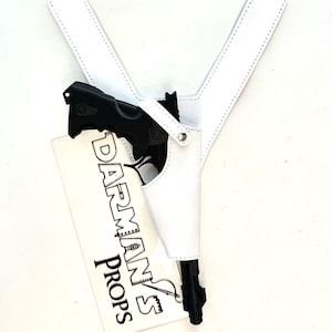 May include: A white leather holster with a black handgun. The holster is in a Y-shape with a stitched border. A white tag with the text "DARMAN'S PROPS" is visible.
