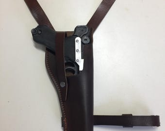 May include: A brown leather holster with a black gun inside. The holster has a strap that goes over the shoulder and another strap that goes around the leg.