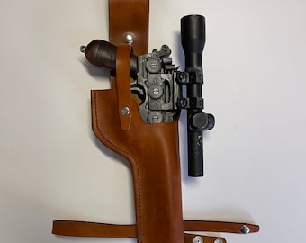 May include: A brown leather holster with a black scope attached to a silver pistol. The holster has a strap that can be attached to a belt.
