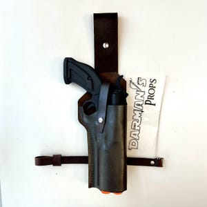 May include: A dark brown leather holster containing a black prop pistol, featuring a vertical belt strap with a silver metal rivet and a horizontal leg strap. A white rectangular sticker with the black text DARMAN'S PROPS is positioned nearby.