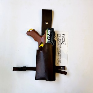 May include: A brown leather holster with a black strap and a black and green toy gun with the word "RIVAL" printed on it. The holster is attached to a white wall with a white paper label that says "DARMAN'S PROPS".