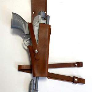 Könnte beinhalten: Ein braunes Lederholster mit einer silberfarbenen Pistole darin. Das Holster hat einen Riemen, der um die Taille geht.