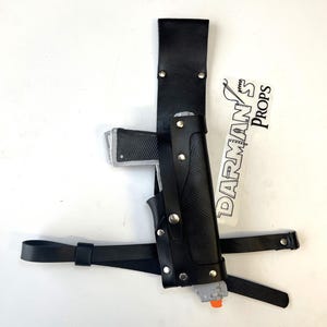 May include: A black leather holster with a silver and black toy gun. The holster has a strap that goes around the waist and a second strap that goes over the shoulder. The holster is labeled "DARMAN'S PROPS".