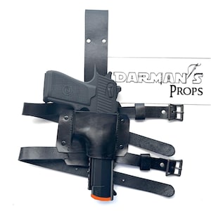 May include: Black leather holster with a black handgun and a black strap. The holster is designed to be worn on the thigh. The strap is adjustable and has a buckle. The holster is labeled "Darman's Props".